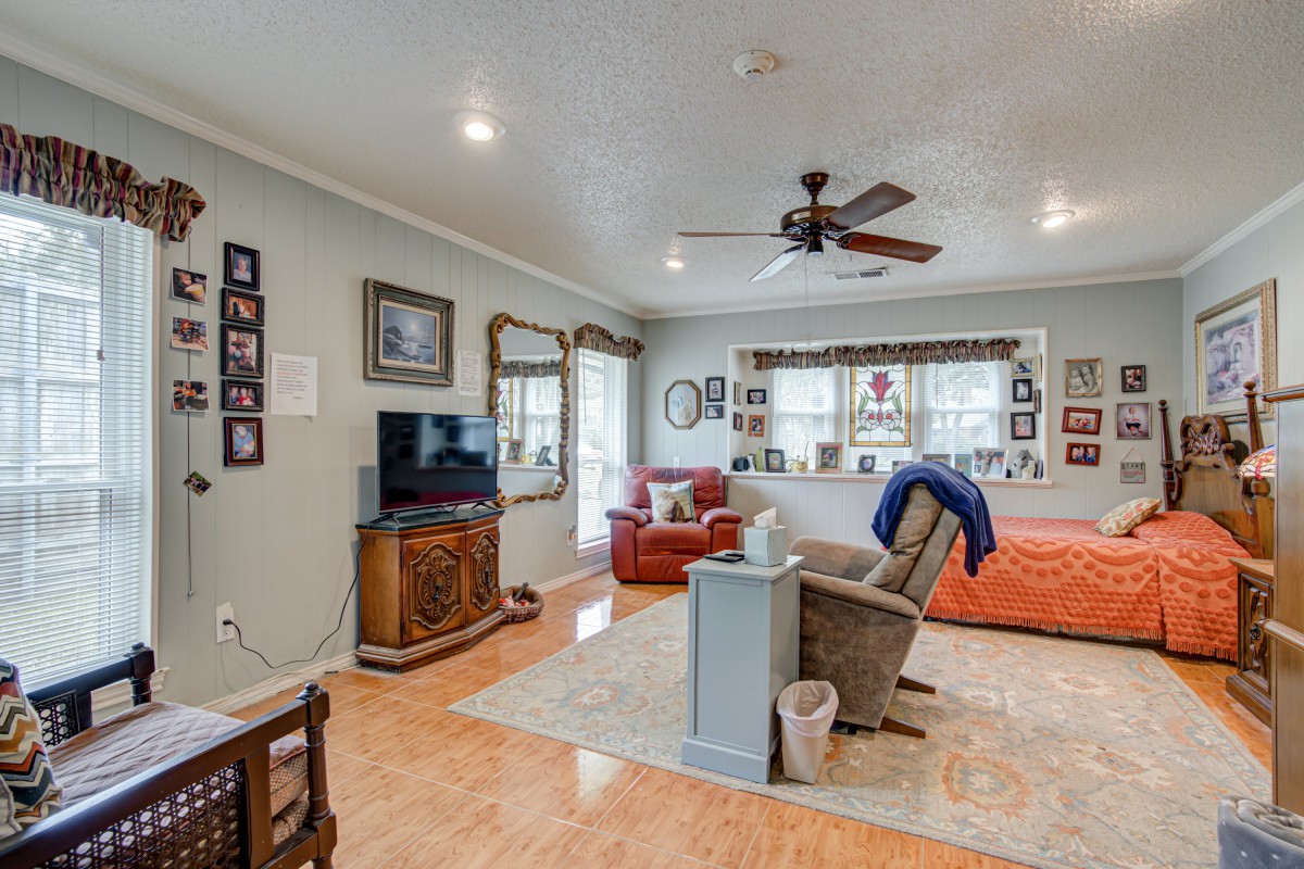 A cozy living room with a comfortable arrangement of furniture, including a sofa, chair, and television. The walls are adorned with various framed photos, and the room features hardwood flooring and a ceiling fan.