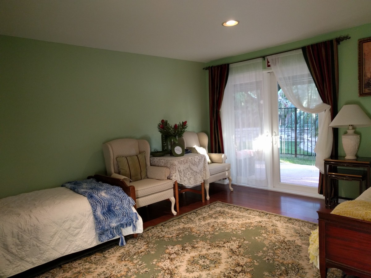 A cozy bedroom with a bed, two armchairs, and a rug, illuminated by natural light through sheer curtains.