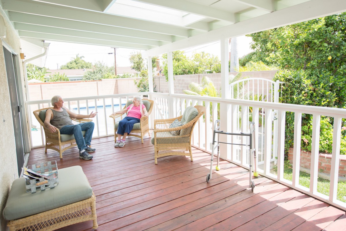 Elderly couple sitting on a porch with wicker furniture and a walker nearby.