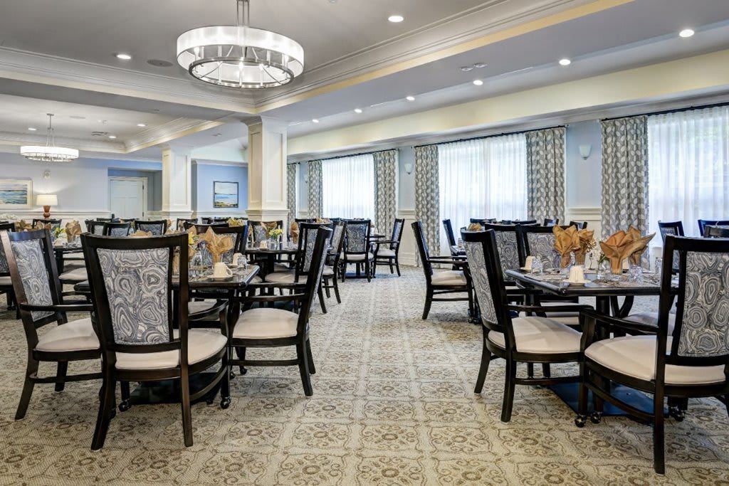 An elegant dining room with neatly arranged tables and chairs, featuring a chandelier and large windows.