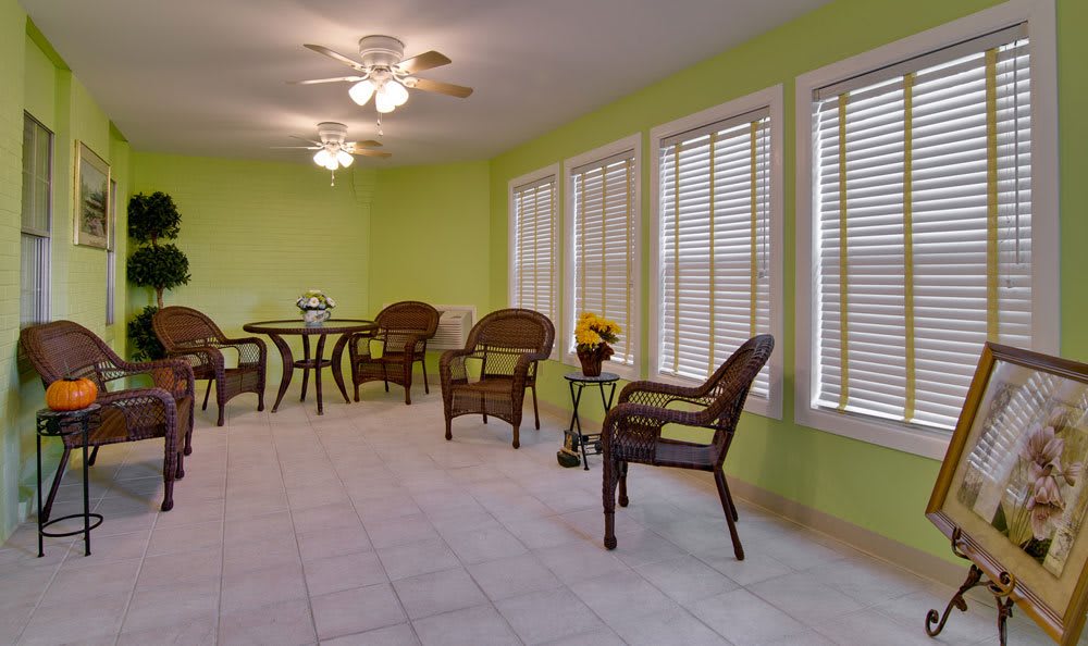 A bright sunroom with rattan furniture, large windows with blinds, and a ceiling fan.