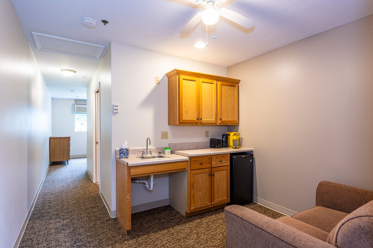 A compact kitchenette in a studio apartment featuring wooden cabinets, a small refrigerator, and essential appliances.