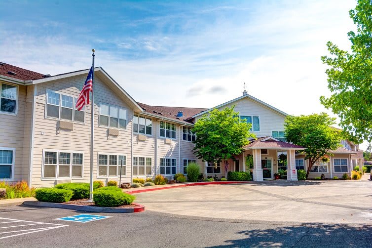A two-story residential building with an American flag in front, surrounded by trees and a parking area.