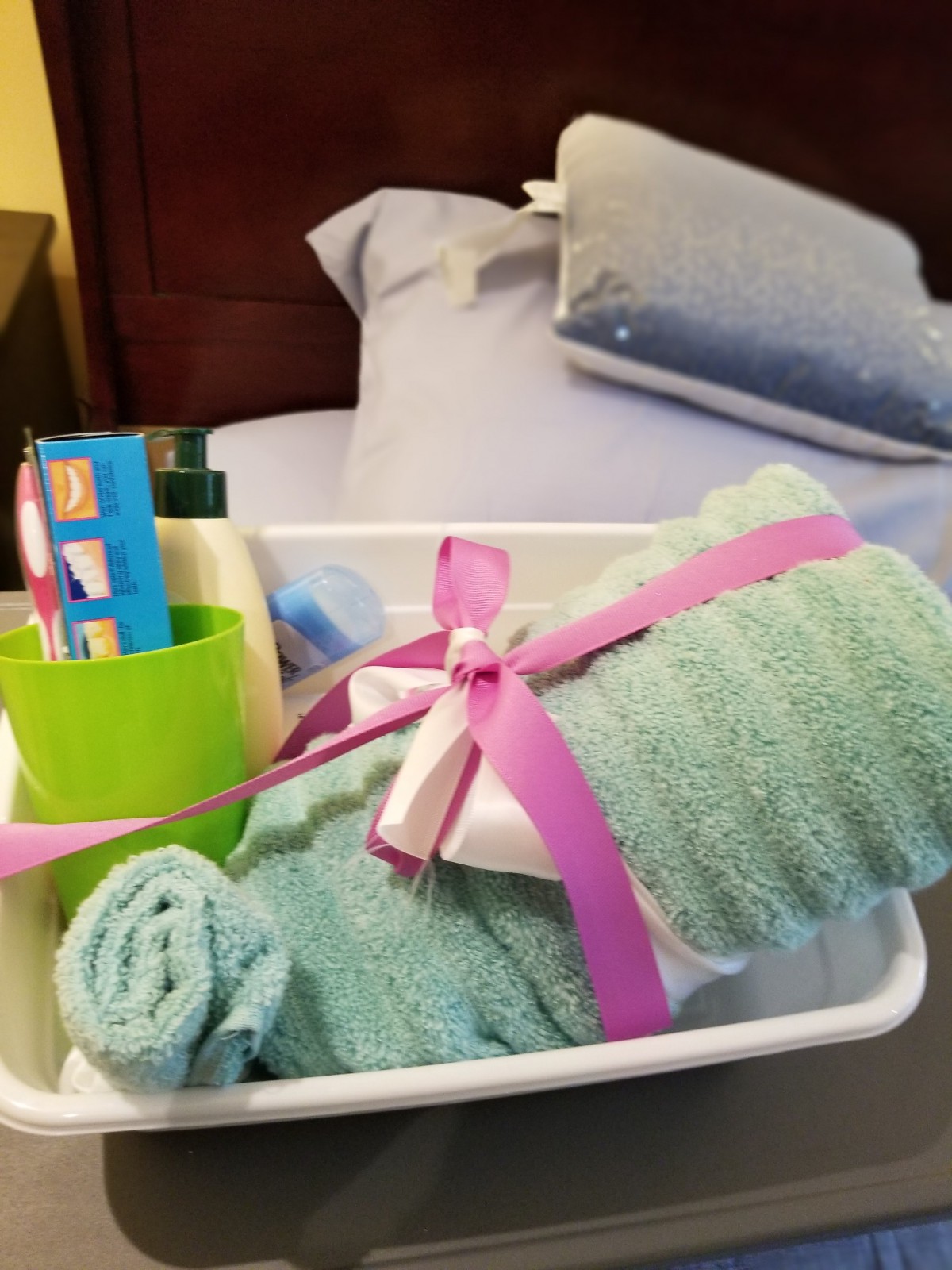 A gift basket containing towels and bathroom essentials with a pink ribbon, placed on a bed.