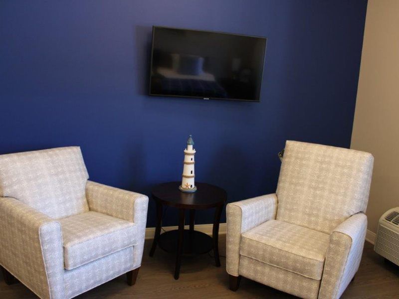 Two patterned chairs beside a small side table with a lighthouse ornament, situated under a wall-mounted television against a blue wall.