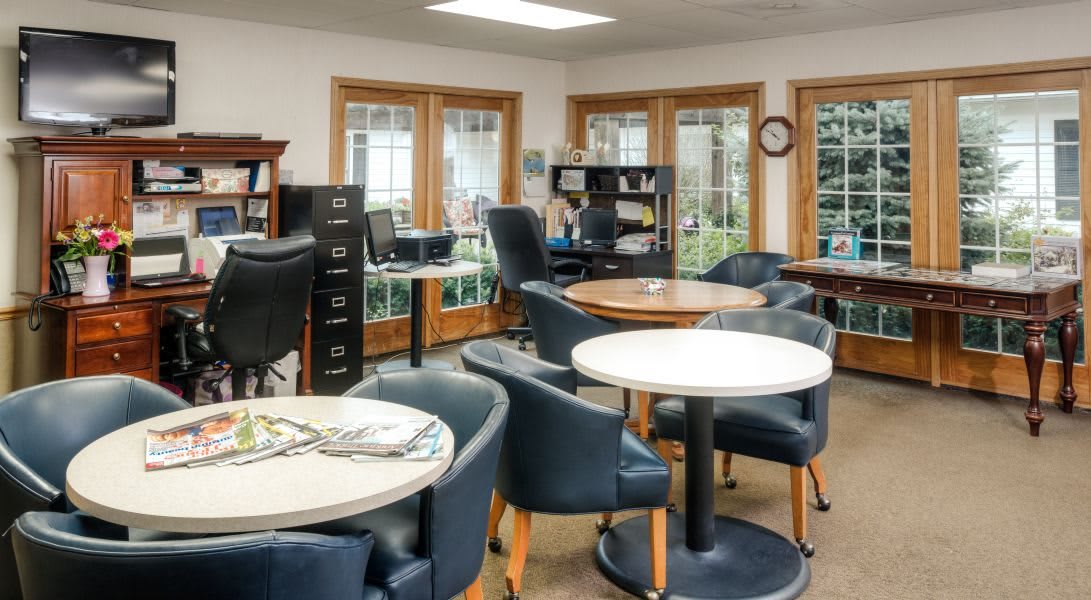 A well-lit office space with tables, chairs, and bookshelves near large windows.