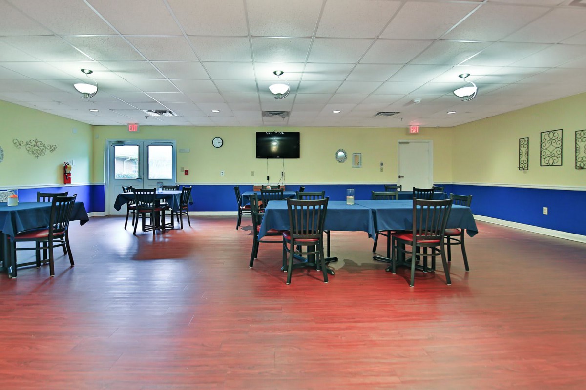 An empty dining room with several tables and chairs arranged, featuring a wooden floor and a wall-mounted television.