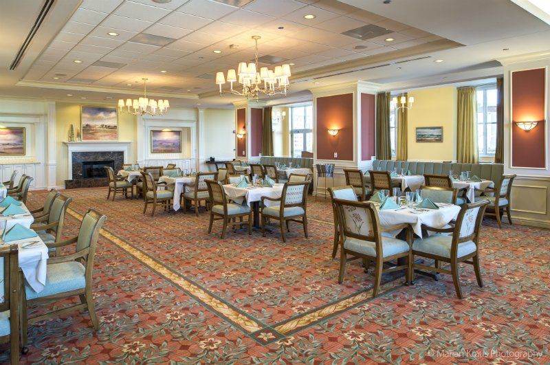 An elegant dining room with neatly arranged tables and chairs, featuring chandeliers and a carpeted floor.