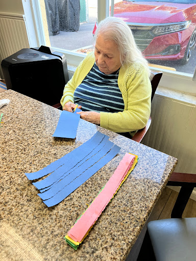 An elderly woman engages in a crafting activity, working with blue and pink paper strips at a table.