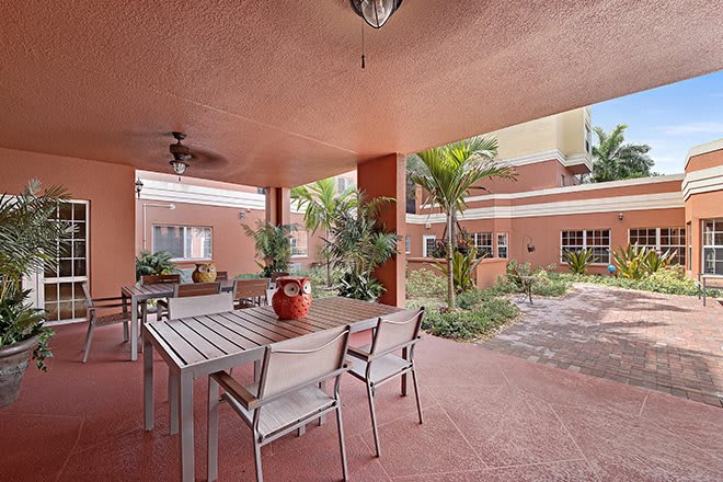 A covered outdoor patio with a table and chairs, surrounded by plants and part of a building's exterior.