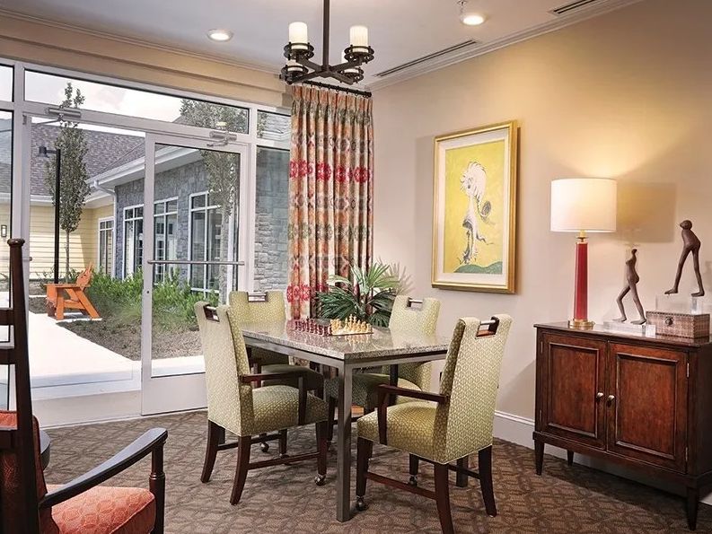 A cozy dining room with a table set for four, featuring artwork on the wall and a large window providing natural light.