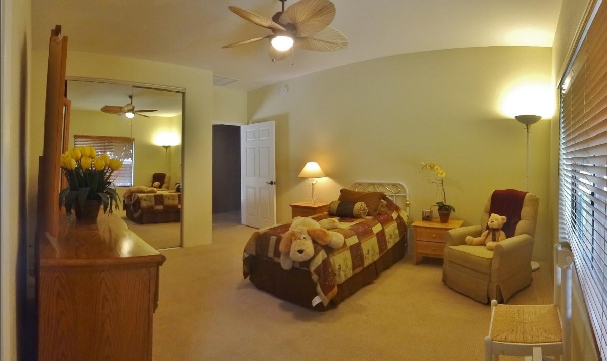 A cozy bedroom with a neatly made bed adorned with a teddy bear, a ceiling fan overhead, and a mirrored closet door reflecting the room.