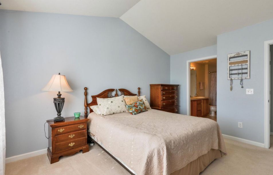 A cozy bedroom with a classic wooden bed, matching nightstand, and dresser. The room features soft blue walls and a lamp providing warm lighting.