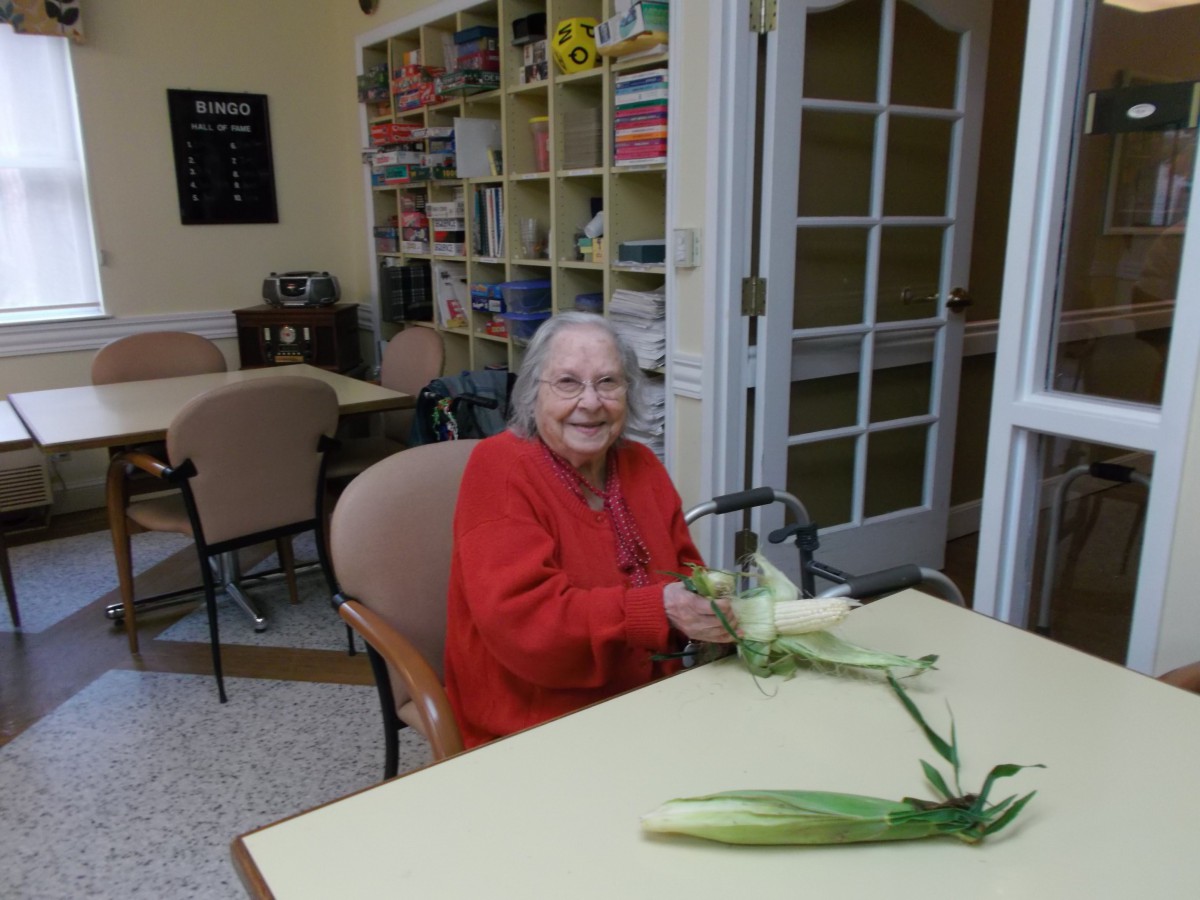An elderly woman sitting at a table, smiling while holding corn in what appears to be a cozy kitchen or dining area.