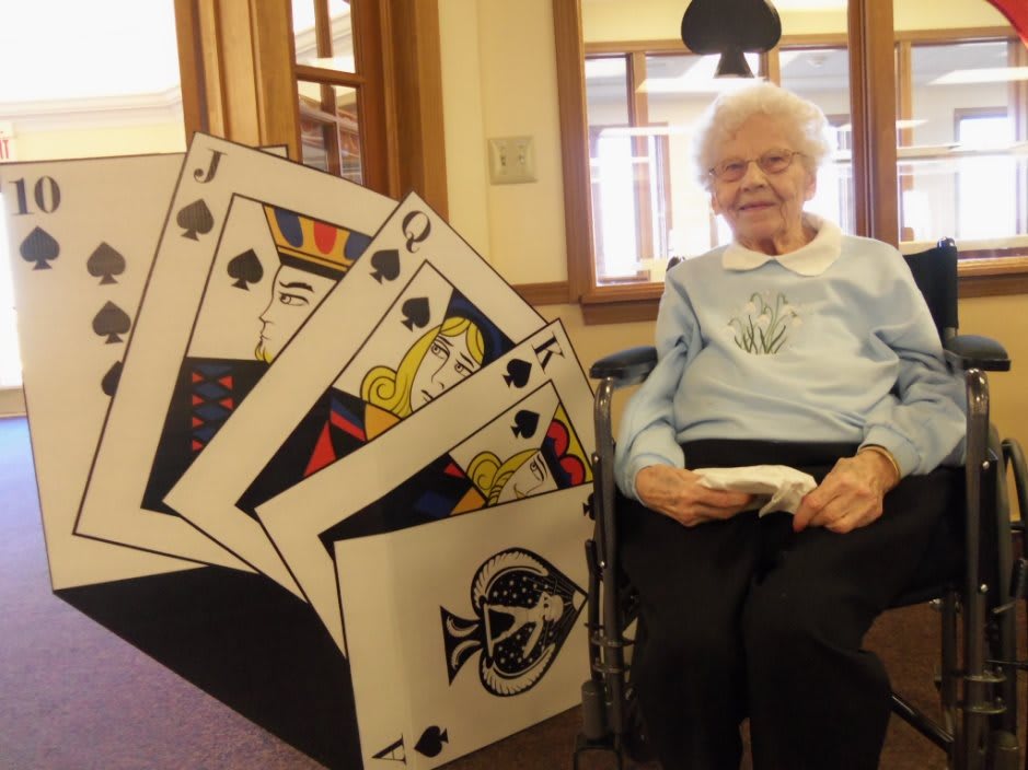 An elderly person sitting indoors next to a display of oversized playing cards.
