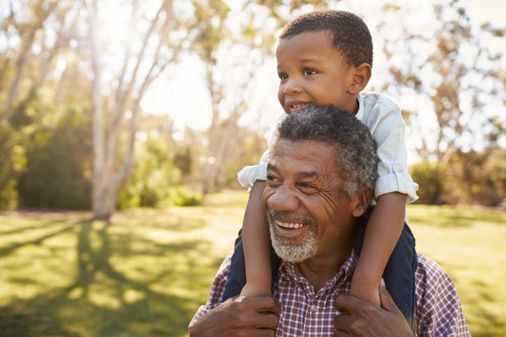 A grandfather carries his smiling grandson on his shoulders in a sunny park.