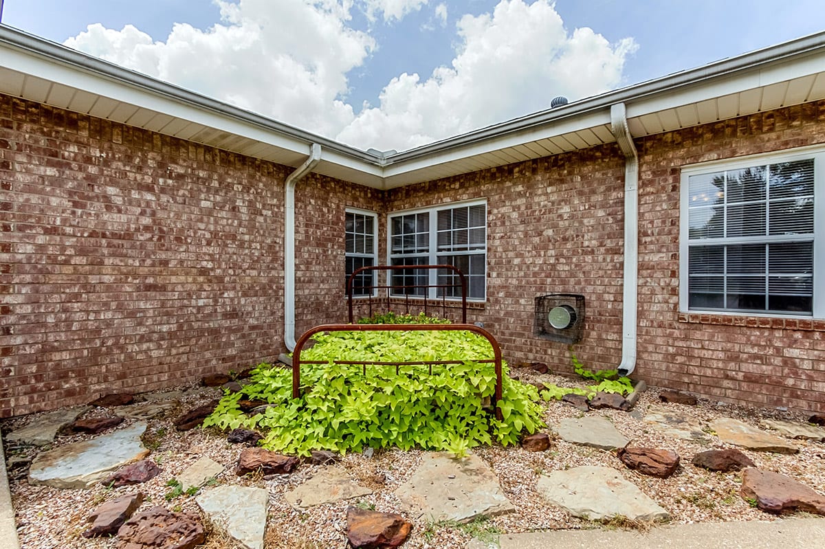 A corner garden with lush greenery and a metal frame against a brick house backdrop.