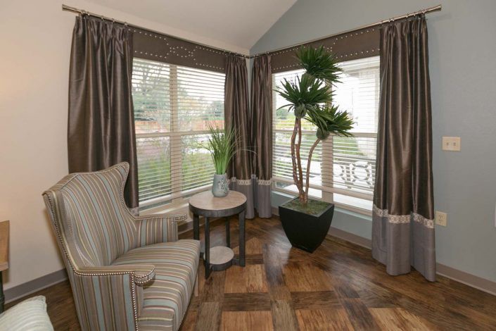 A cozy corner of a living room featuring a striped armchair, large potted plant, and windows with curtains.