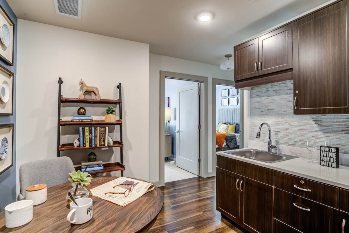 A modern kitchen and dining area with wooden floors, an open door leading to another room, and a shelf with books and decor.
