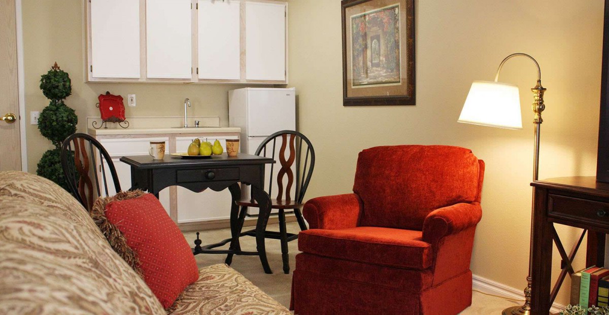A cozy living space with a red armchair, a small table and chair, and a kitchenette in the background.