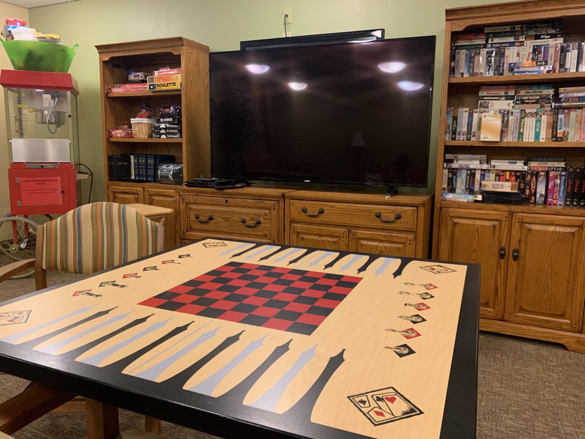 A board game setup on a table in a cozy living room with a large TV and bookshelves in the background.