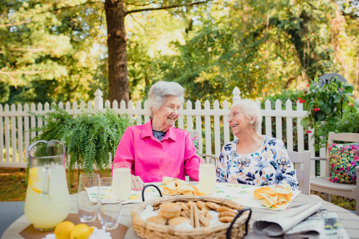 Two elderly women enjoying a conversation over snacks in a garden setting.