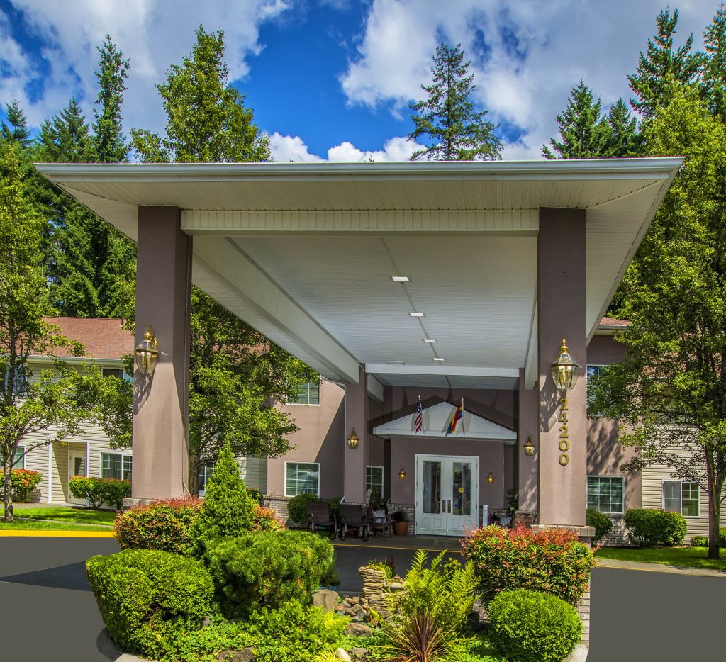 A grand entrance to a building surrounded by lush landscaping and tall trees under a blue sky with clouds.