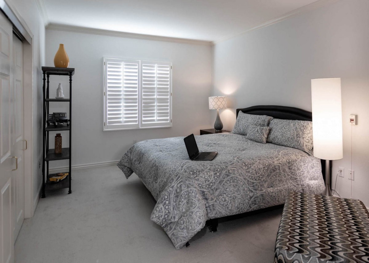 A cozy bedroom with neutral tones, featuring a bed adorned with patterned bedding, a laptop, a lamp, and a bookshelf near the window with shutters.