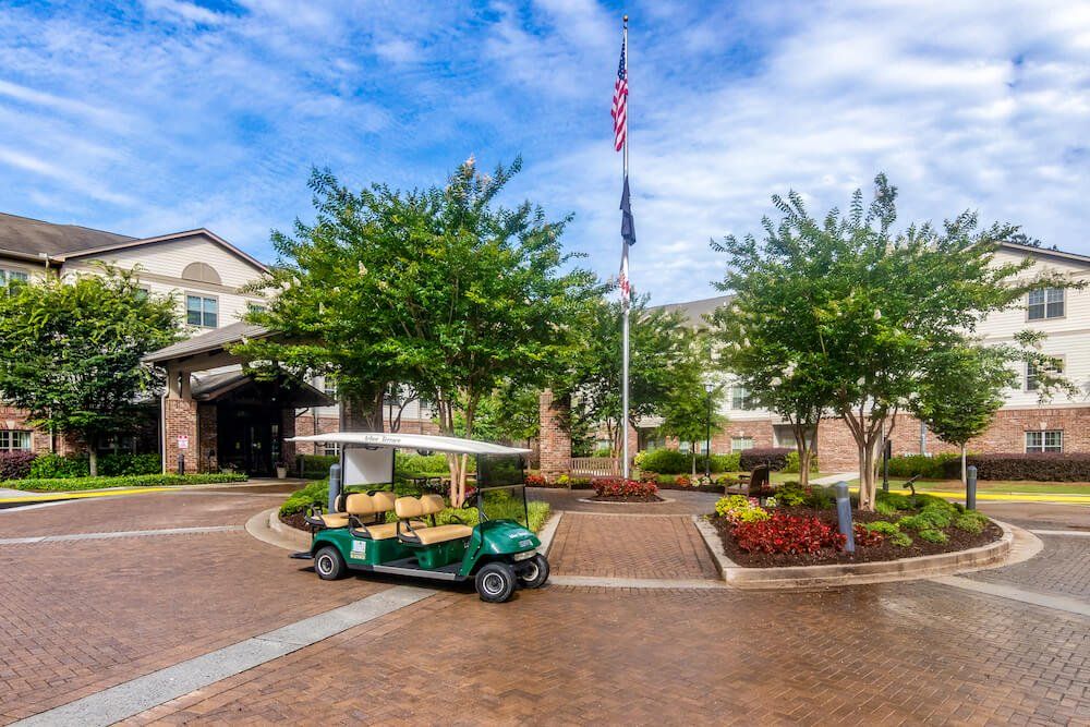 A green golf cart is parked in front of a landscaped building with an American flag flying on a flagpole under a partly cloudy sky.