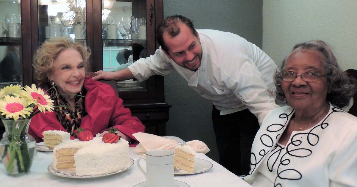 A chef is smiling and interacting with an elderly woman at a table with cake during a celebration.
