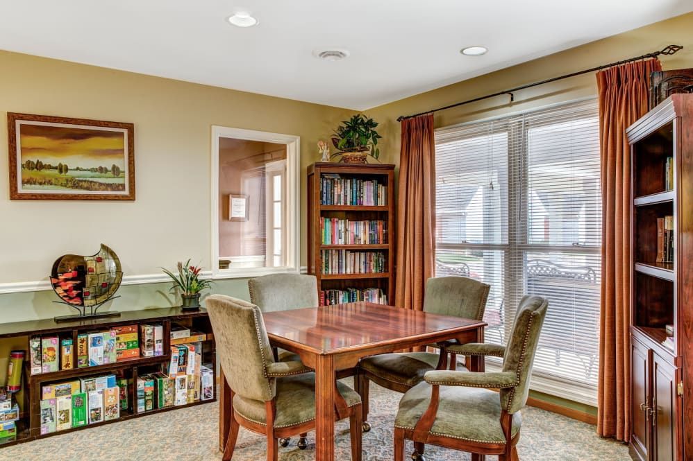 A cozy reading room with a wooden table, chairs, and a well-stocked bookshelf near large windows.