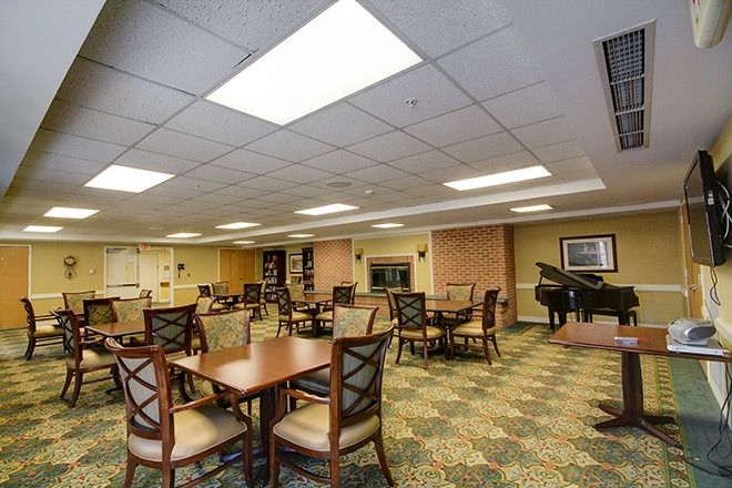 A spacious dining room with multiple tables and chairs, featuring a piano in the corner and overhead ceiling lights.