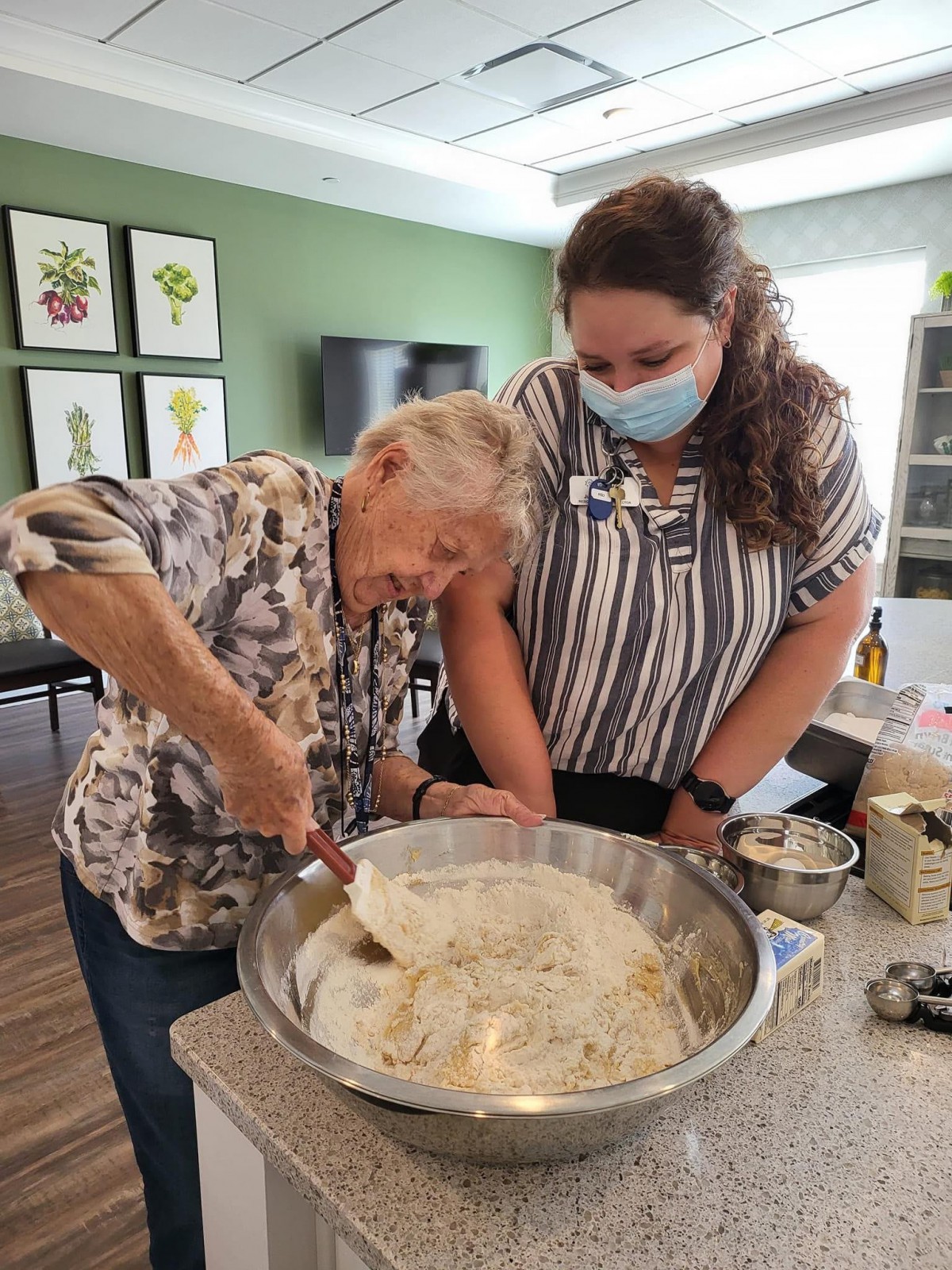 An elderly woman and a young woman working together in the kitchen, mixing ingredients in a large bowl.
