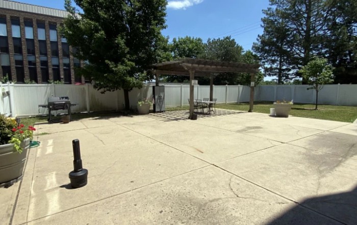 A sunny patio area with a pergola, surrounded by trees and a white fence.