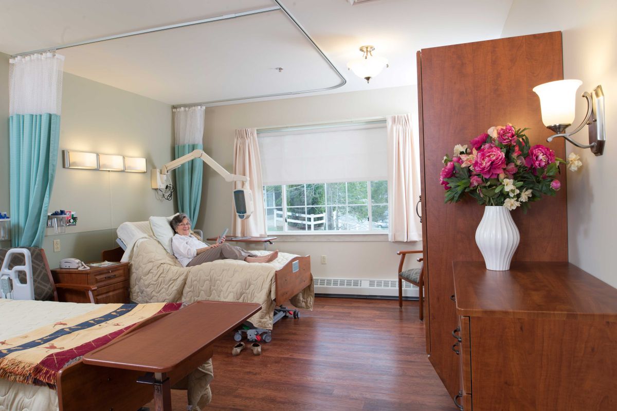 A hospital room with a patient in bed, featuring a large window and a vase of flowers.