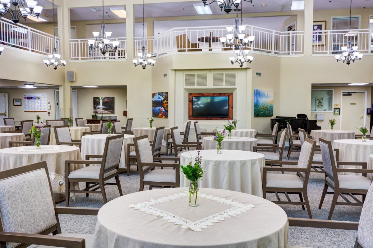 A spacious and elegantly decorated dining area featuring round tables set with white cloths and simple centerpieces, under subtle chandelier lighting.