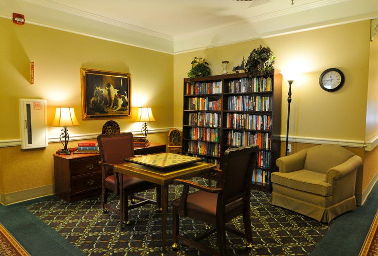 A cozy reading room with bookshelves, a table and chairs, warmly lit by lamps.