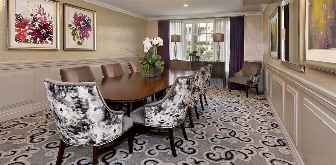 A sophisticated dining room with a large wooden table surrounded by patterned chairs, elegant decor, and ample natural light from the windows.
