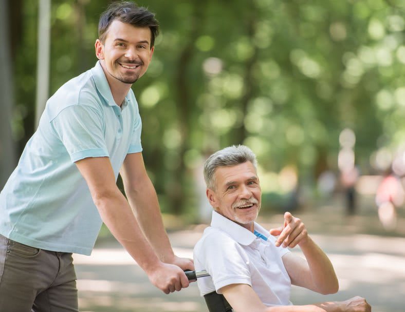 A caregiver pushing an elderly man in a wheelchair through a park.