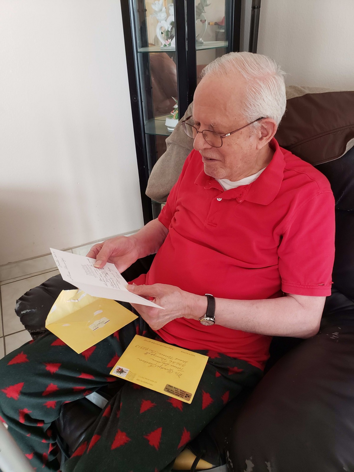 An elderly man in a red shirt is sitting and reading a letter indoors.