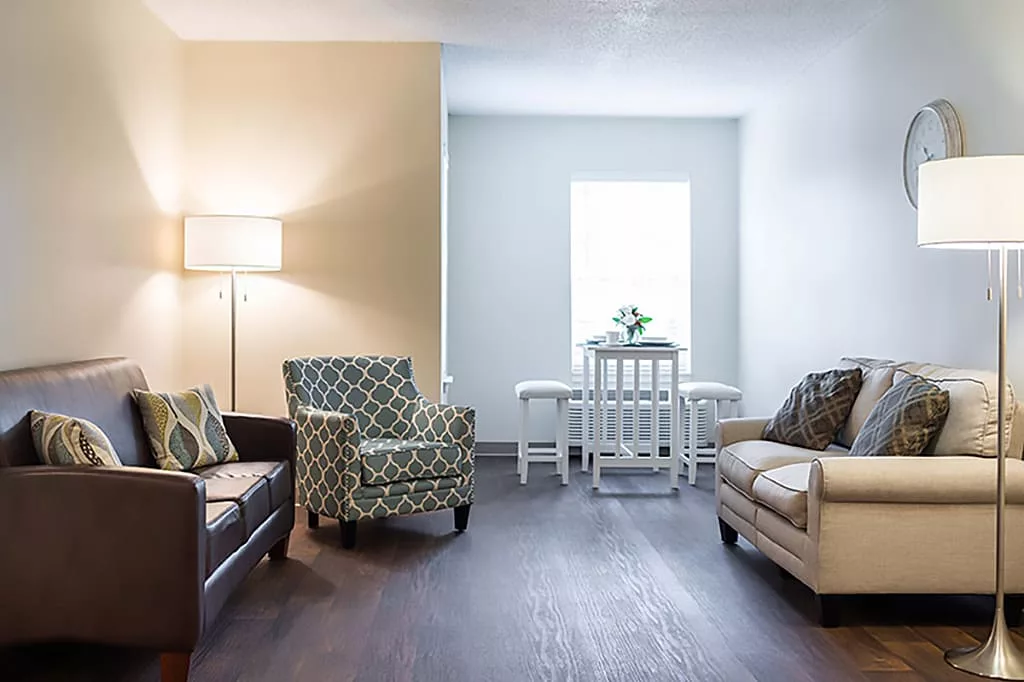 A cozy living room with modern furniture, featuring a patterned armchair, sofa, lamp, and a small dining area by the window.