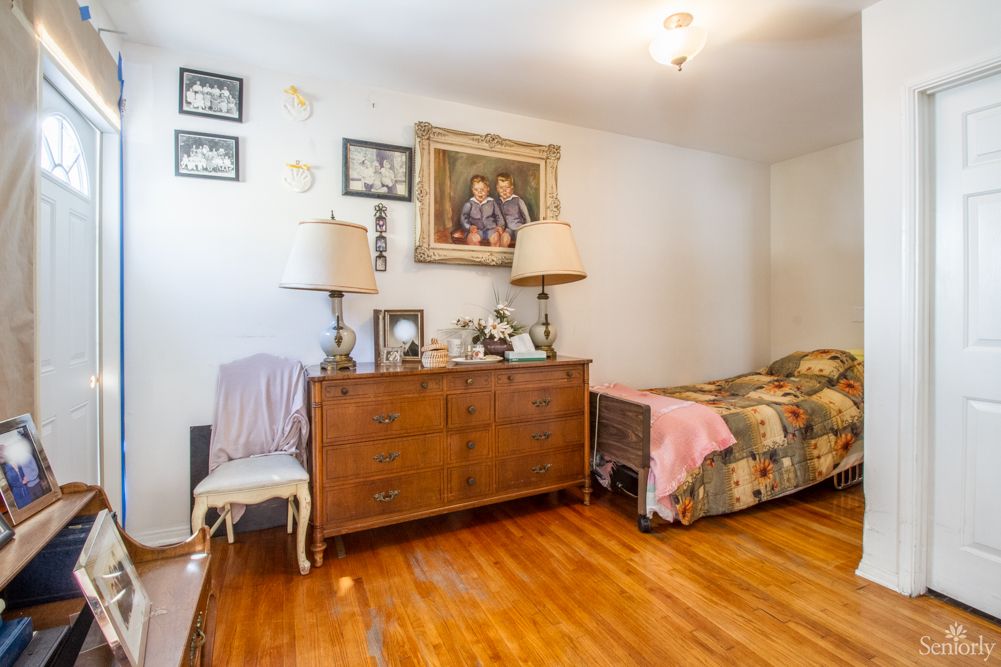 A cozy bedroom featuring a wooden dresser with two lamps and wall art, alongside a single bed with a colorful quilt on a hardwood floor.