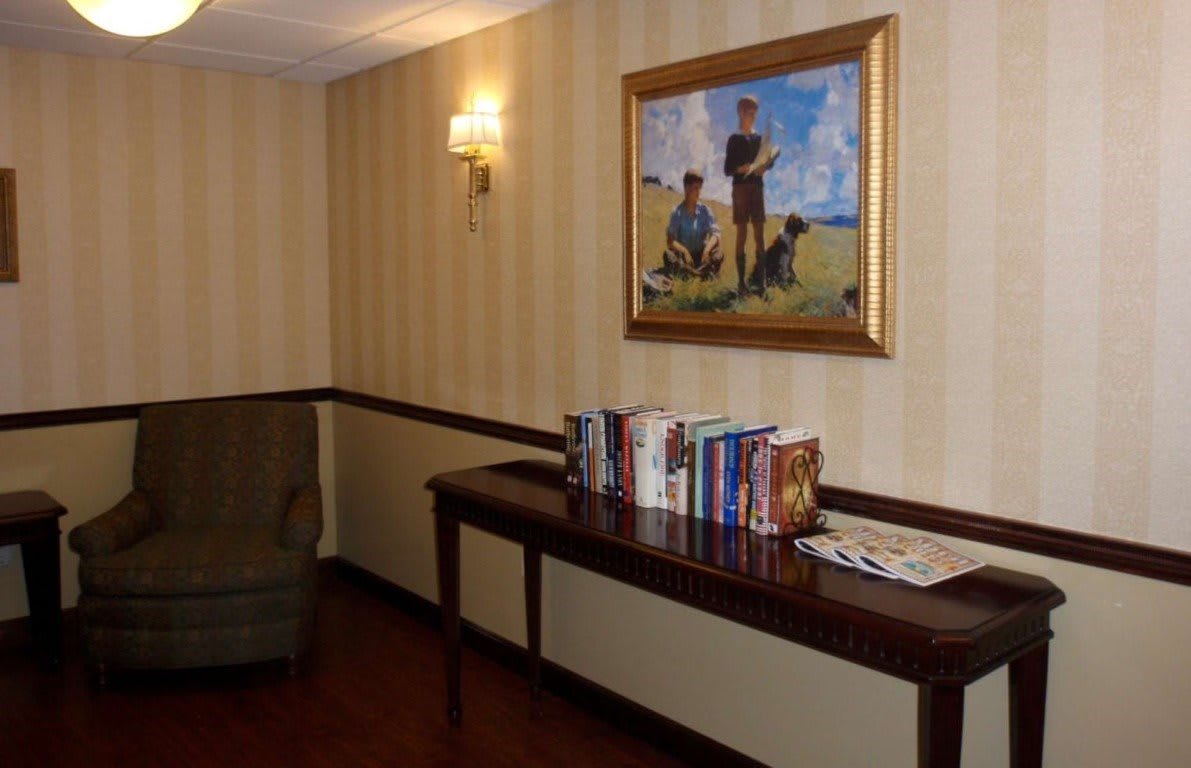 A cozy corner with a wooden table displaying books beneath a framed painting, illuminated by a wall sconce against striped wallpaper.