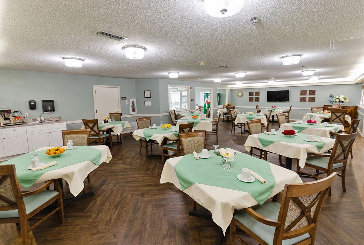 A well-lit dining room with multiple tables set for guests, featuring green and white tablecloths.