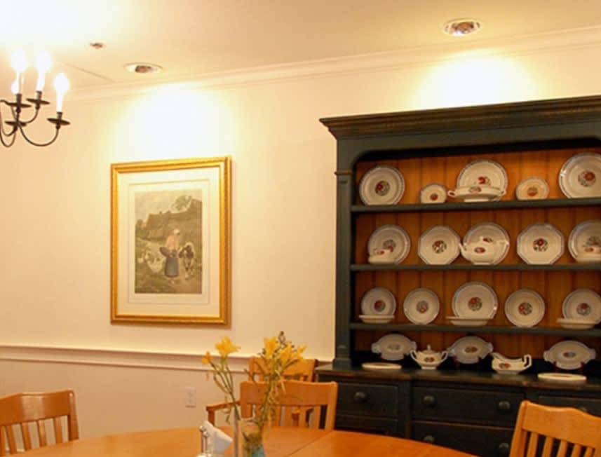 A cozy dining room featuring a china cabinet filled with plates and a wooden table adorned with flowers, complemented by framed artwork on the wall.