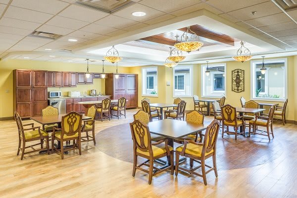 A well-lit dining area with wooden tables and chairs, featuring chandeliers and an open kitchen space.