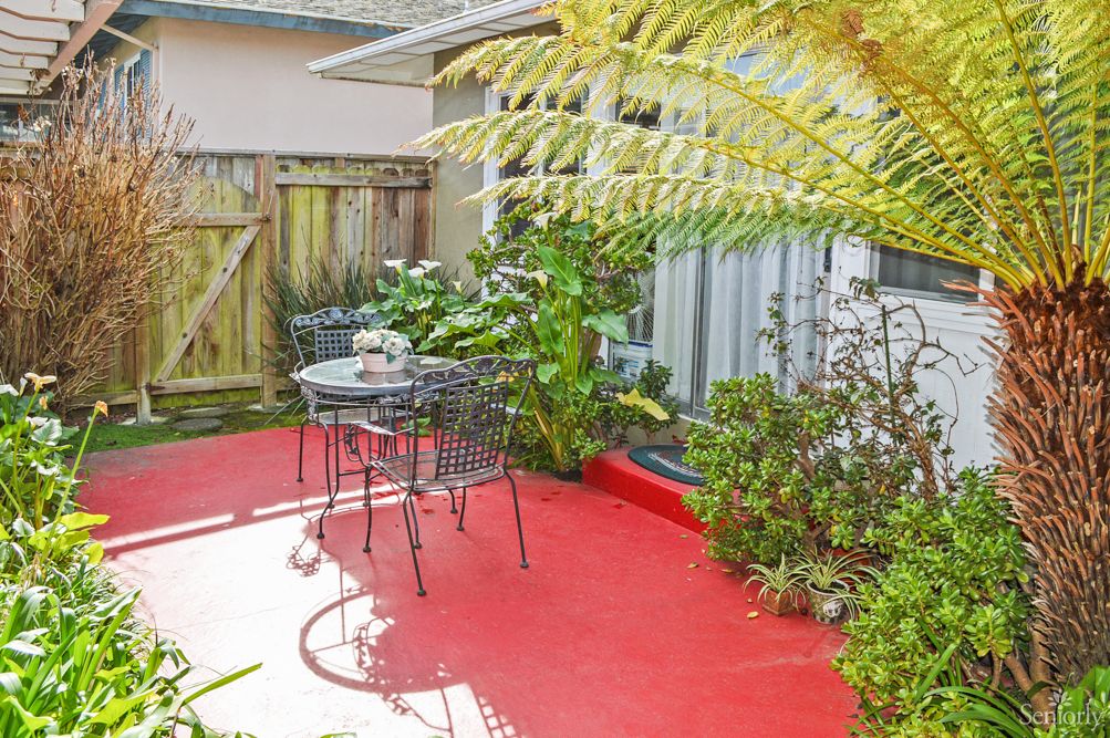 A cozy patio with a red floor, surrounded by lush plants and featuring a small outdoor table and chairs.