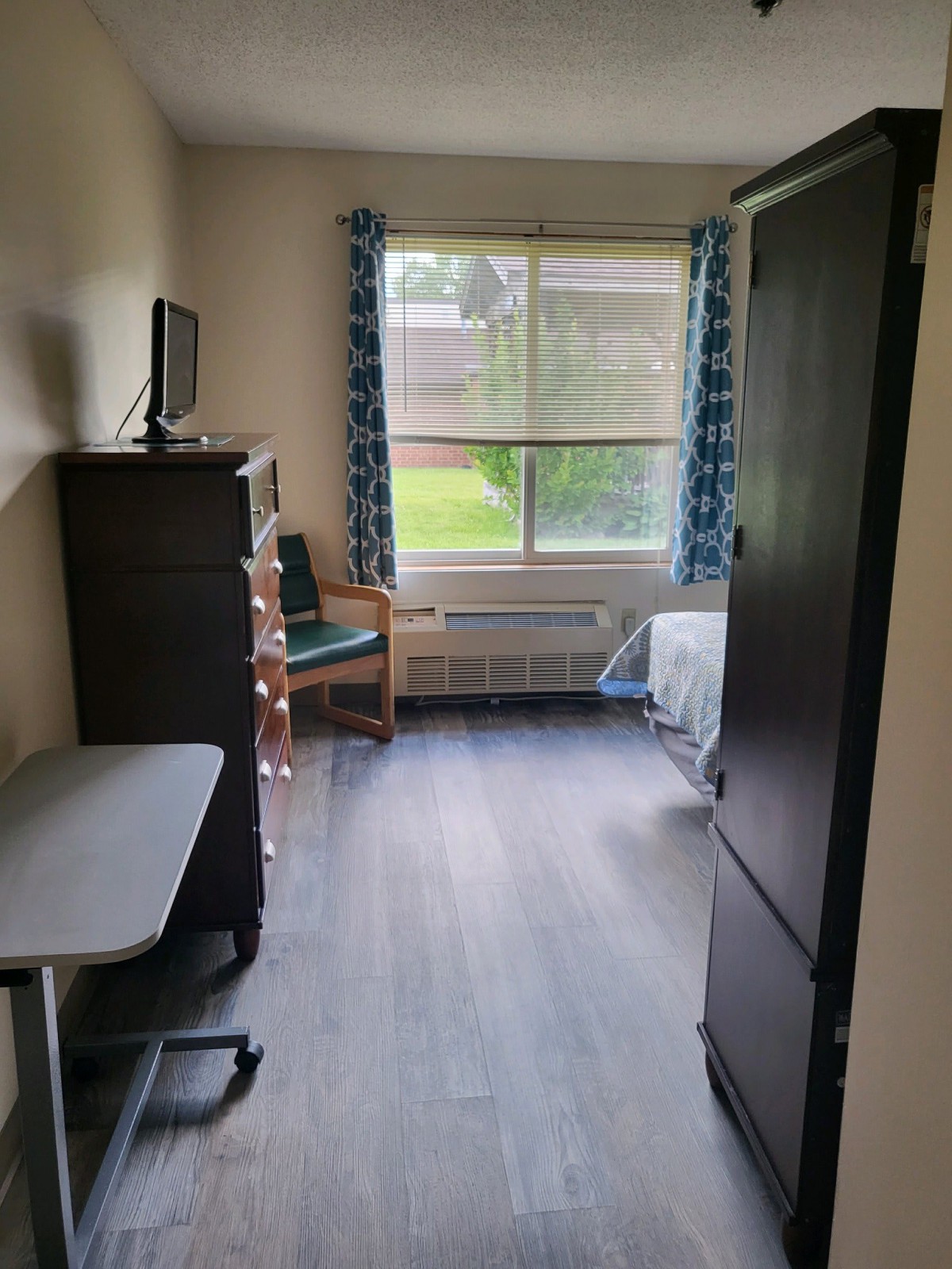 A cozy dorm room with a bed, desk, wardrobe, and television under a large window.