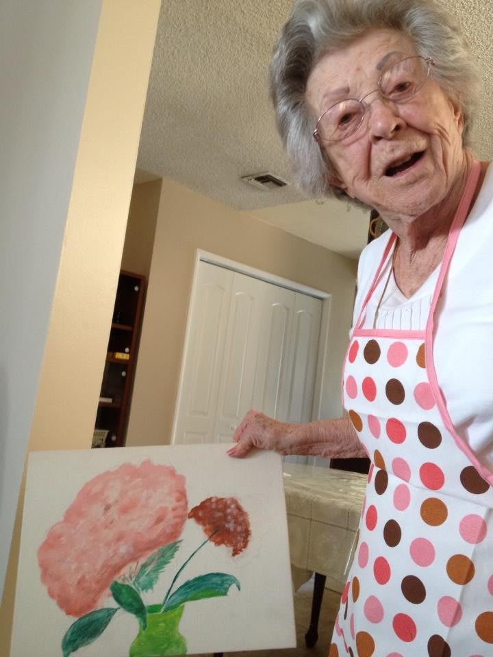 An elderly woman wearing a polka dot apron proudly displays her flower painting indoors.