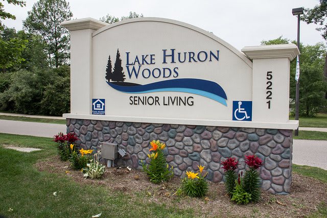 Entrance sign for Lake Huron Woods Senior Living, surrounded by landscaped flowers.